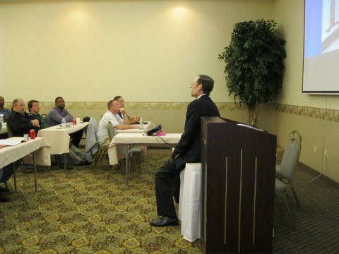 A man is sitting at a podium giving a presentation to a group of people sitting at tables.