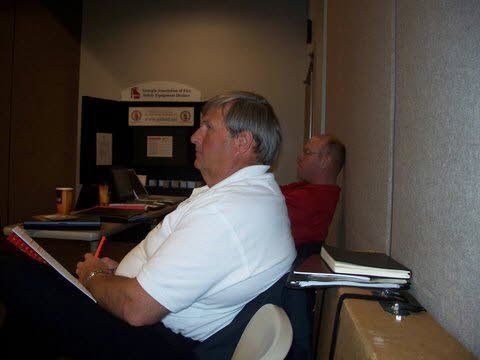 Two men are sitting at a desk in a room.