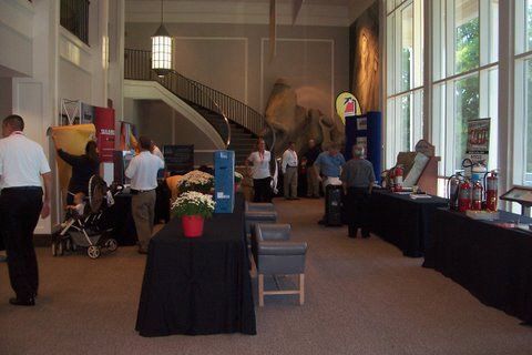 A group of people are standing around tables in a room with a staircase in the background.