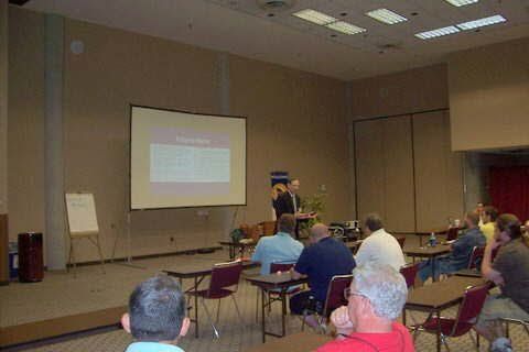 A group of people are sitting at tables in front of a projector screen.