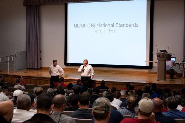 A man is giving a presentation in front of a large screen that says ul/ulc bi-national standards for ul-711