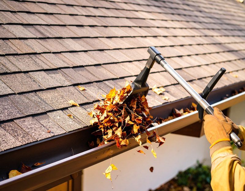 Person cleaning a gutter on a house roof with a long-handled tool, removing leaves and debris.