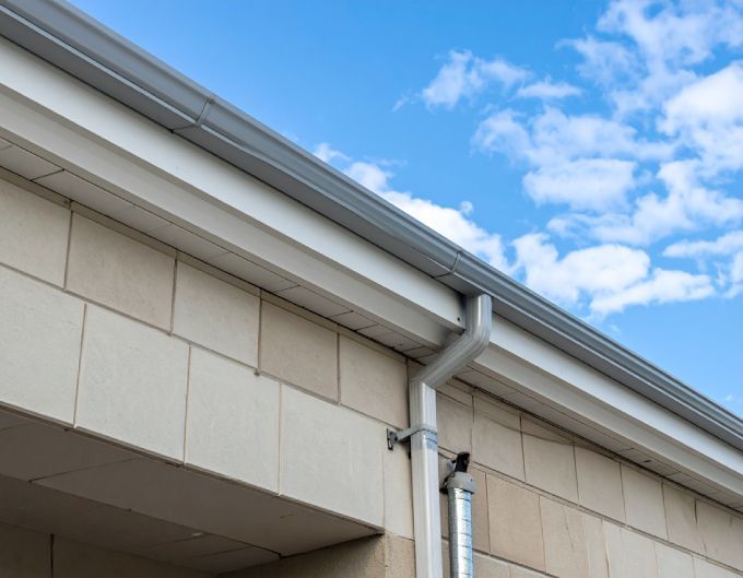 Gray rain gutter attached to a beige brick building, with a blue sky and clouds in the background.