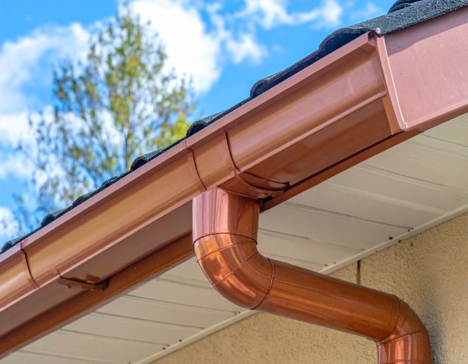 Copper gutters and downspout on a beige house with a blue sky background.