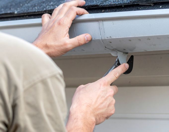 Person installing a gutter, using a tool; hands are visible, working on the edge of a building.