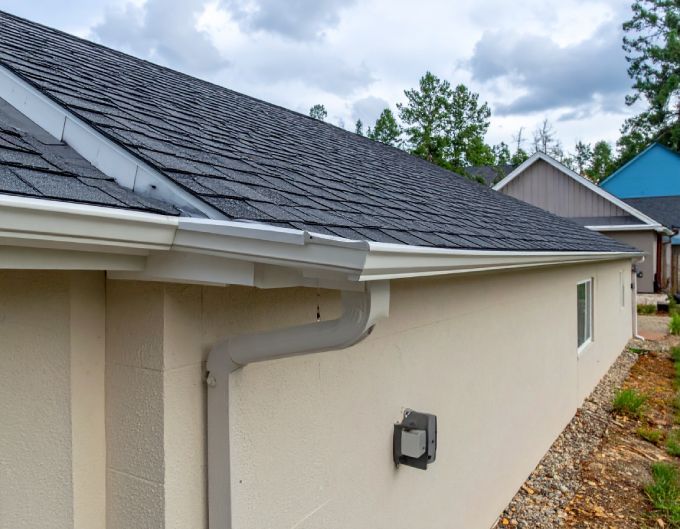 Beige building with dark gray roof and white gutters, cloudy sky background.