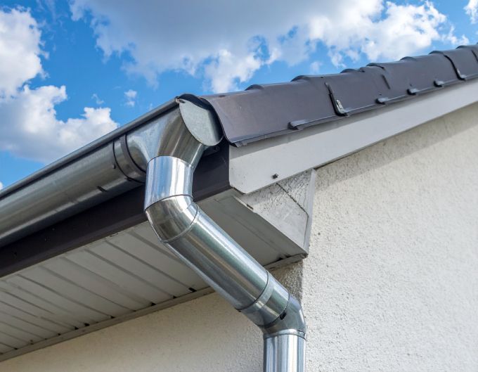 steel gutter and downspout on a white house with a brown roof against a cloudy blue sky.