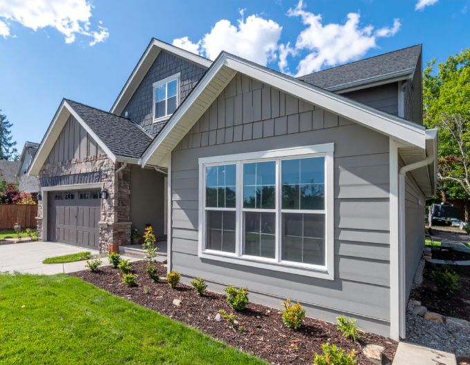 Gray house with white trim and a brown garage, blue sky, and green grass.
