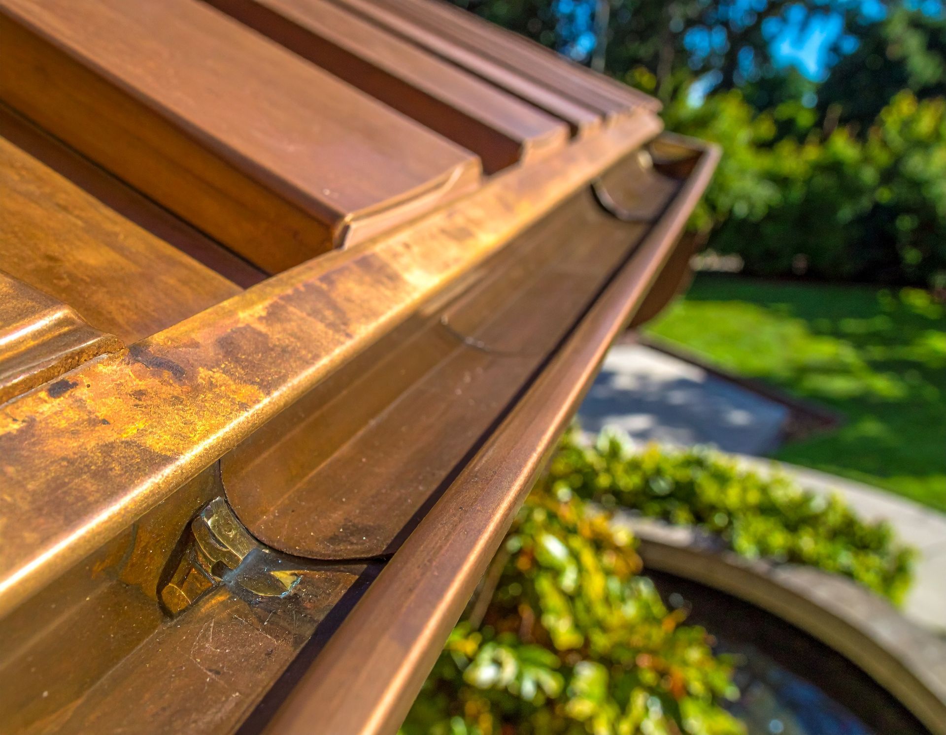 Close-up of a bronze roof gutter and downspout on a sunlit building, with greenery in the background