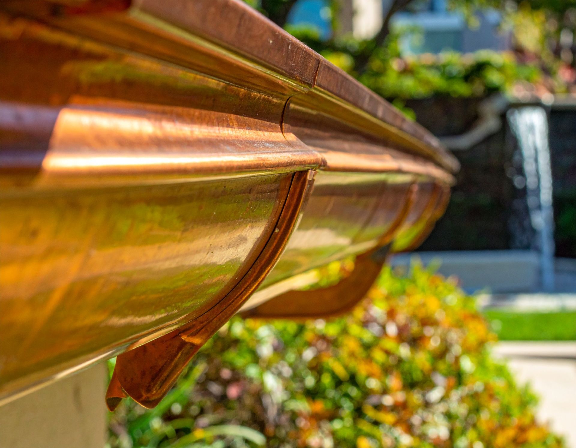 Close-up of a copper gutter on a house roof with sunlit leaves and a blurred garden background