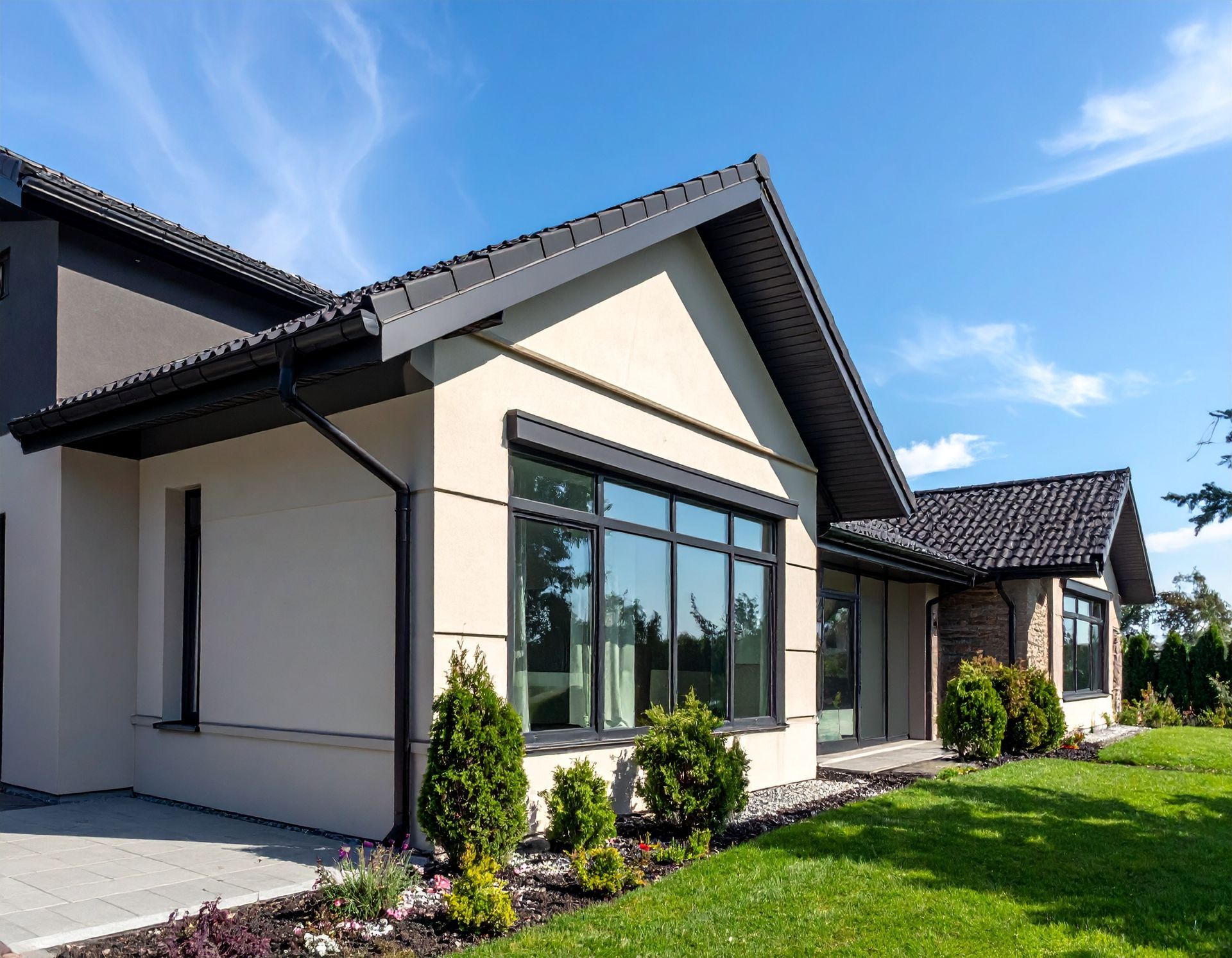 Modern single-story house exterior with white walls, dark roof, large windows, and a green lawn under a blue sky