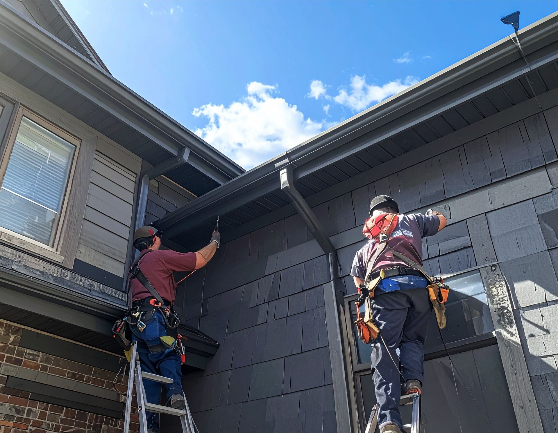 Two roofers on ladders installing dark gray siding on a house exterior under a blue sky.