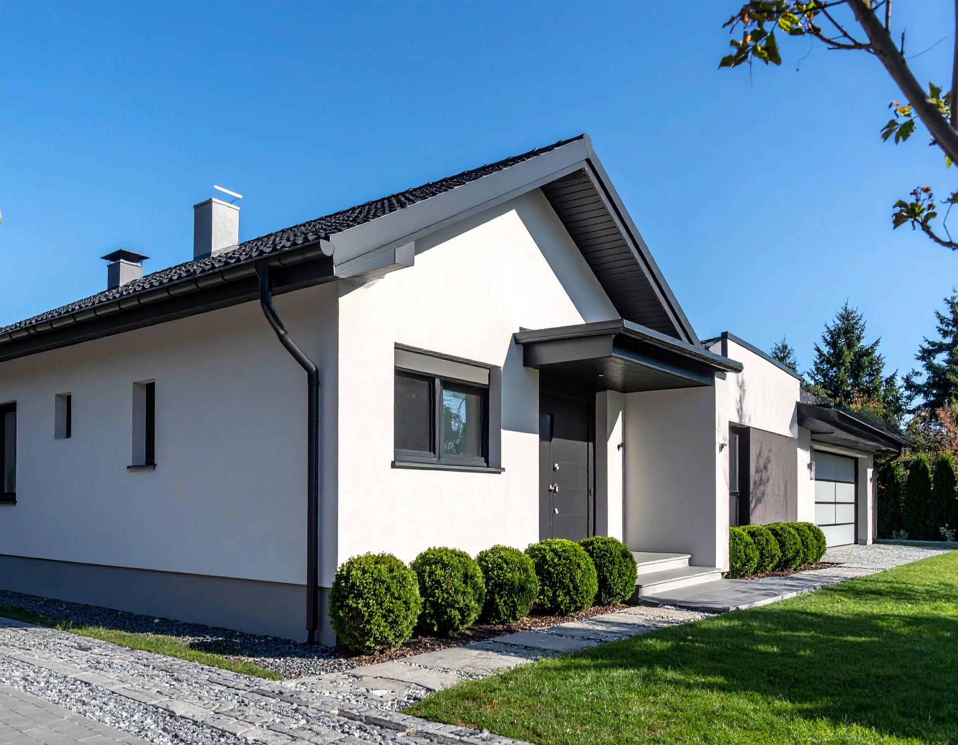 Modern white single-story house with dark roof, small front porch, and neatly trimmed shrubs on a sunny lawn