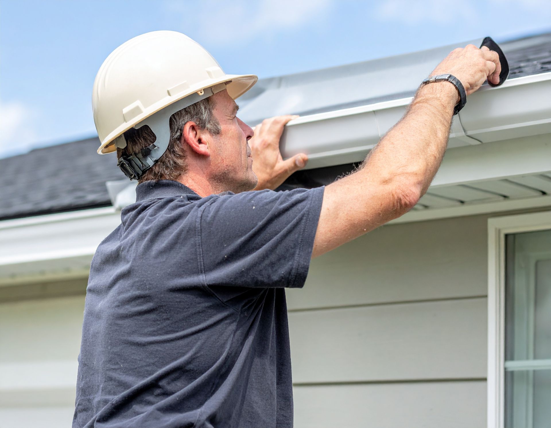 Man in hard hat inspecting white gutter on a house.