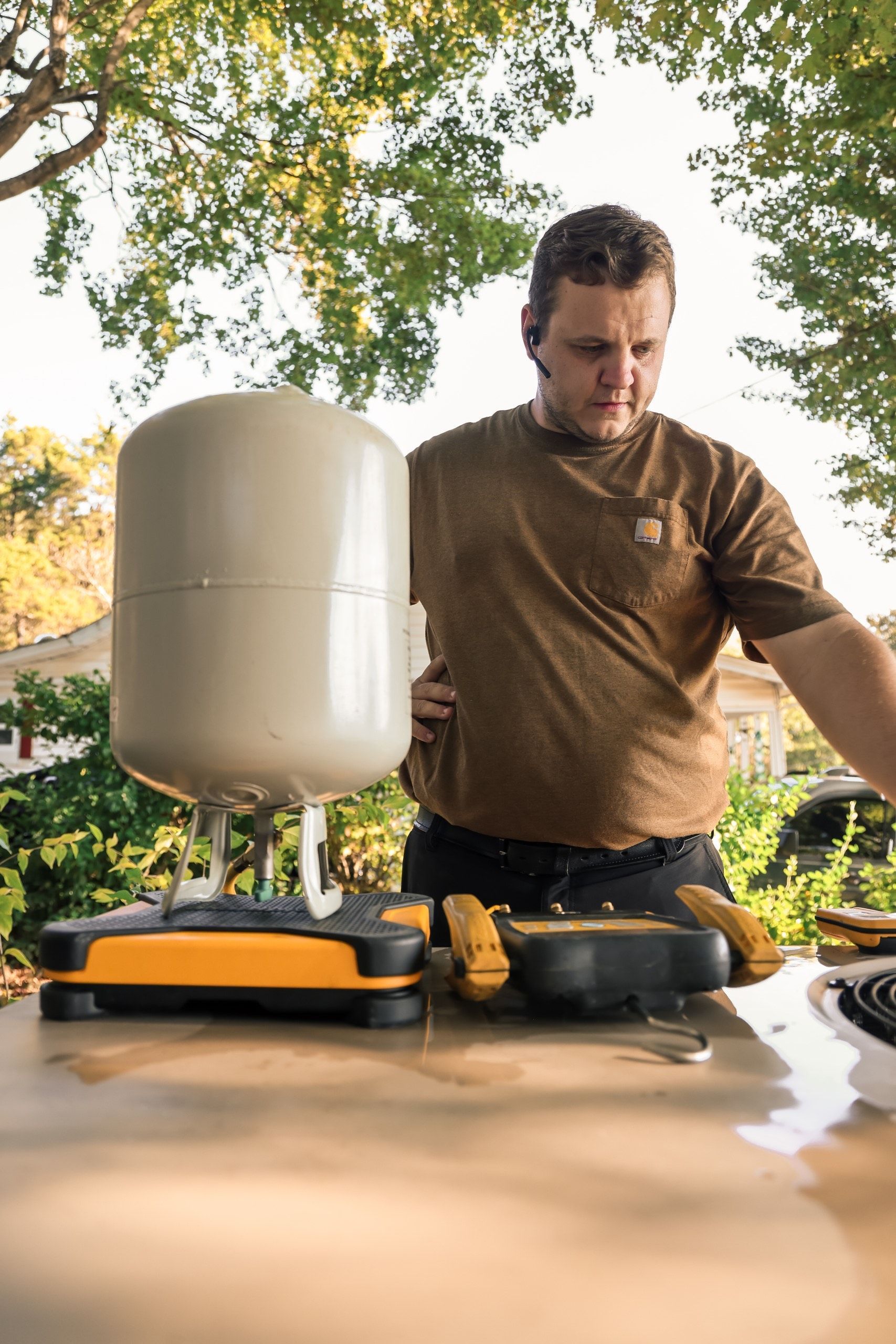 A technician in a brown shirt inspects a refrigerant cylinder and digital gauge resting on an outdoor HVAC unit.