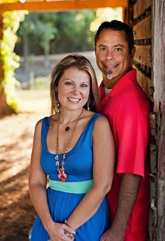 Couple smiling outdoors, woman in blue dress and man in red shirt, leaning against a wooden structure.