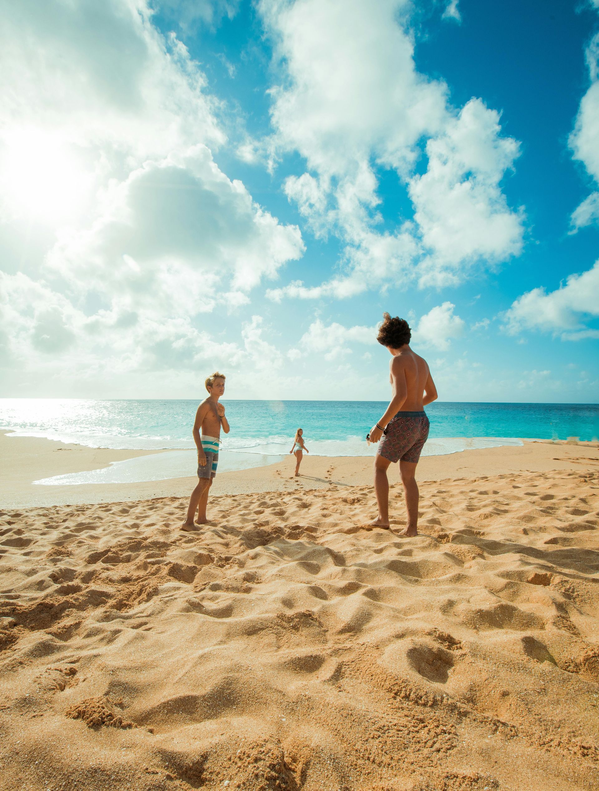 Three people playing on a sunny beach, blue sky with puffy clouds.