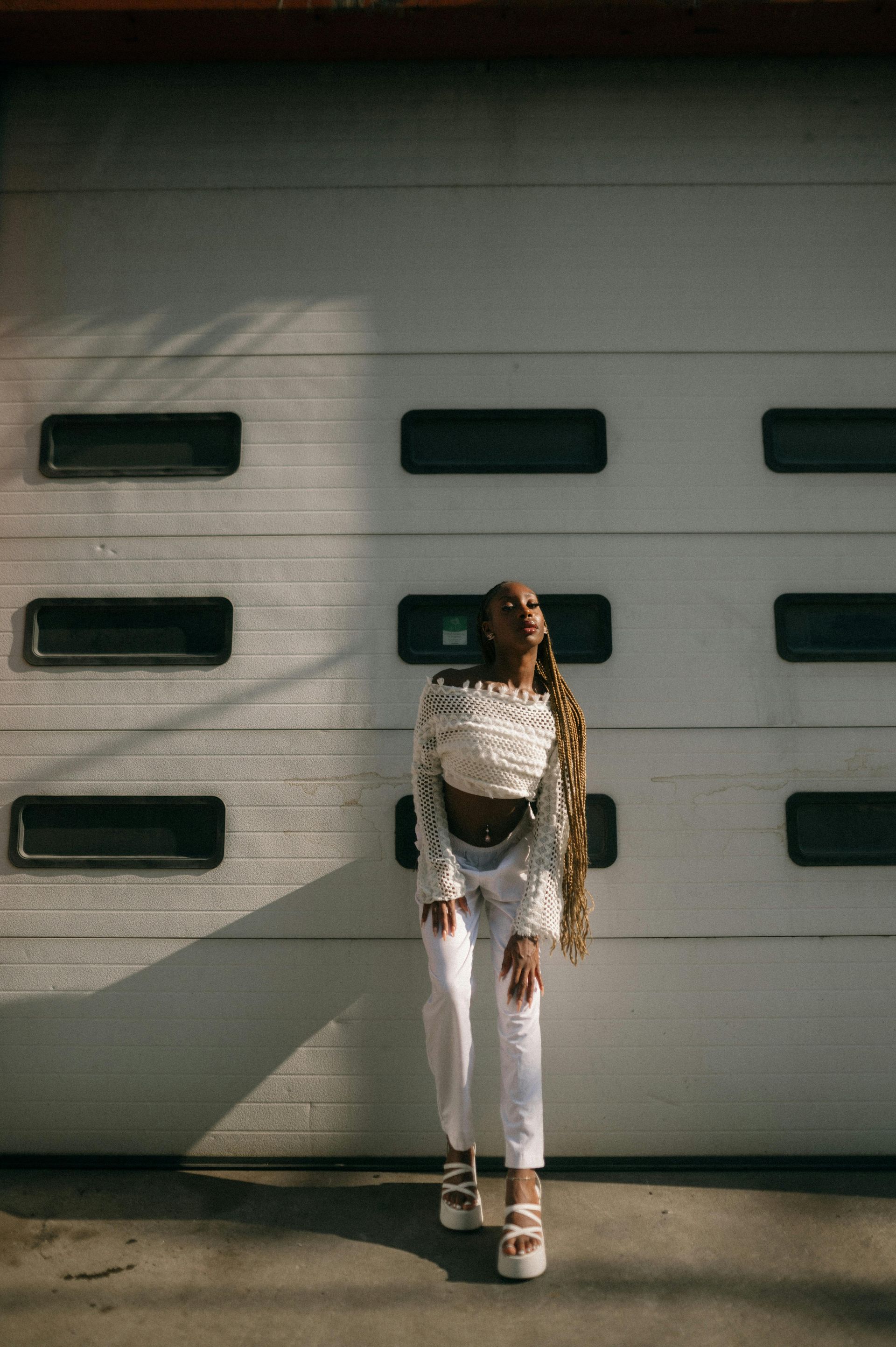 A woman in a white crop top and white jeans is standing in front of a garage door.