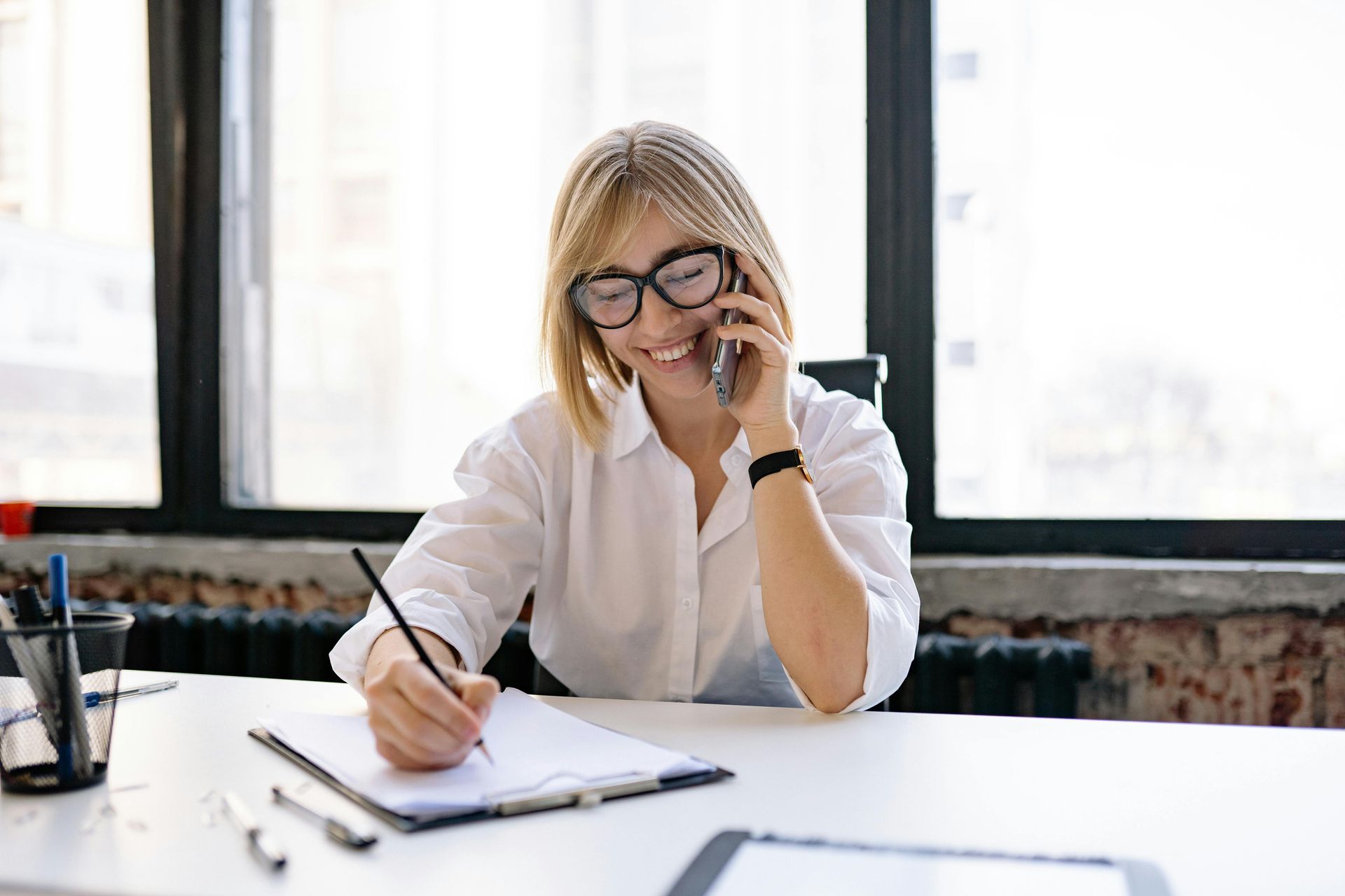 A woman is sitting at a desk talking on a cell phone and writing in a notebook.