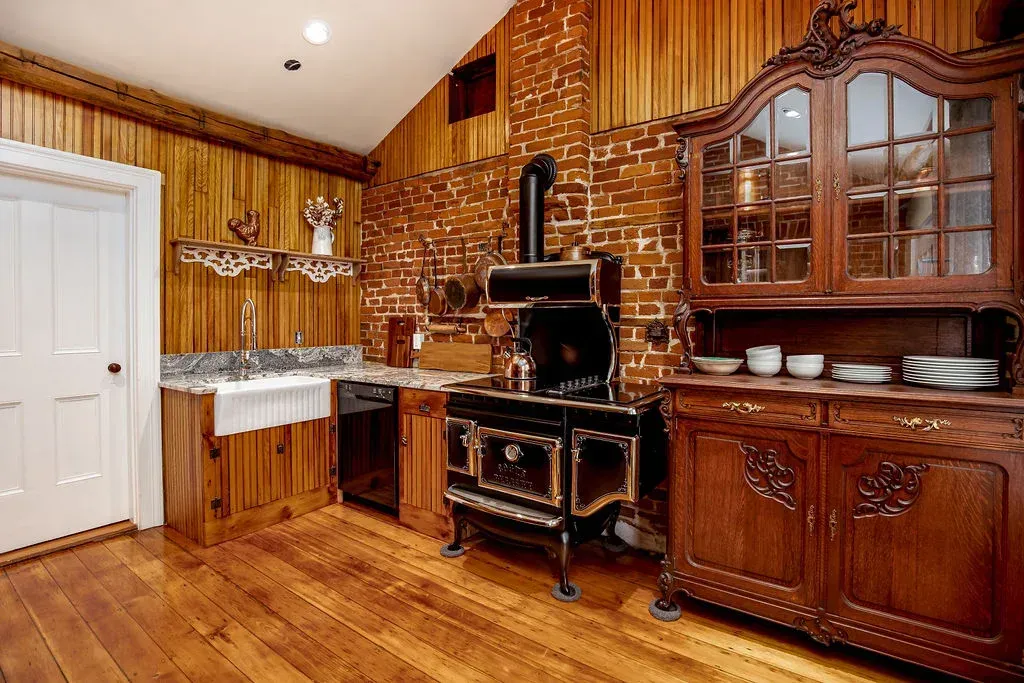 A rustic kitchen features wood-paneled walls, brick accents, a vintage-style stove, a farmhouse sink, and a wooden hutch.