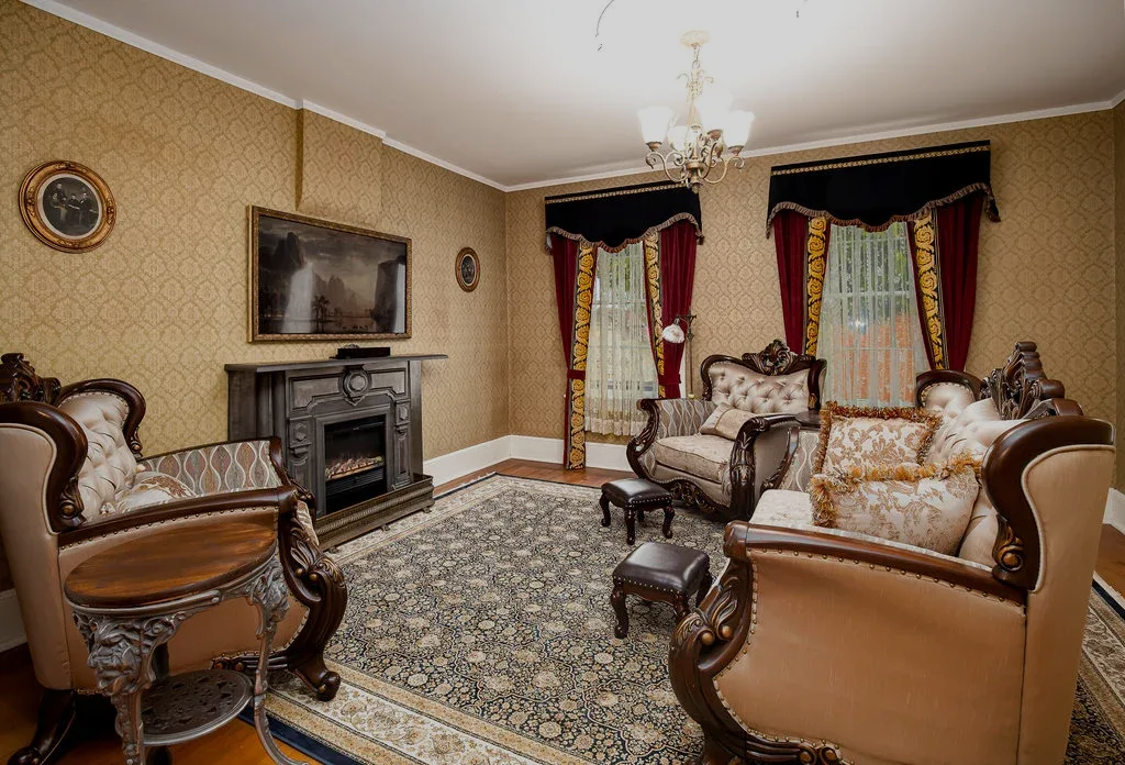 A cozy, traditional living room with patterned wallpaper, ornate wooden armchairs, a fireplace, and a large area rug.
