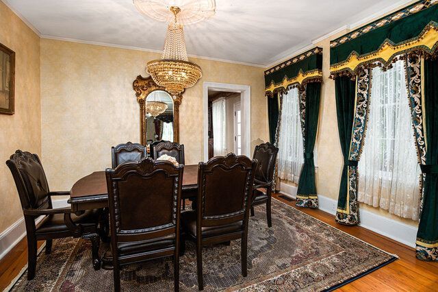 A formal dining room featuring a dark wood table with six matching chairs, a crystal chandelier, and green drapes.