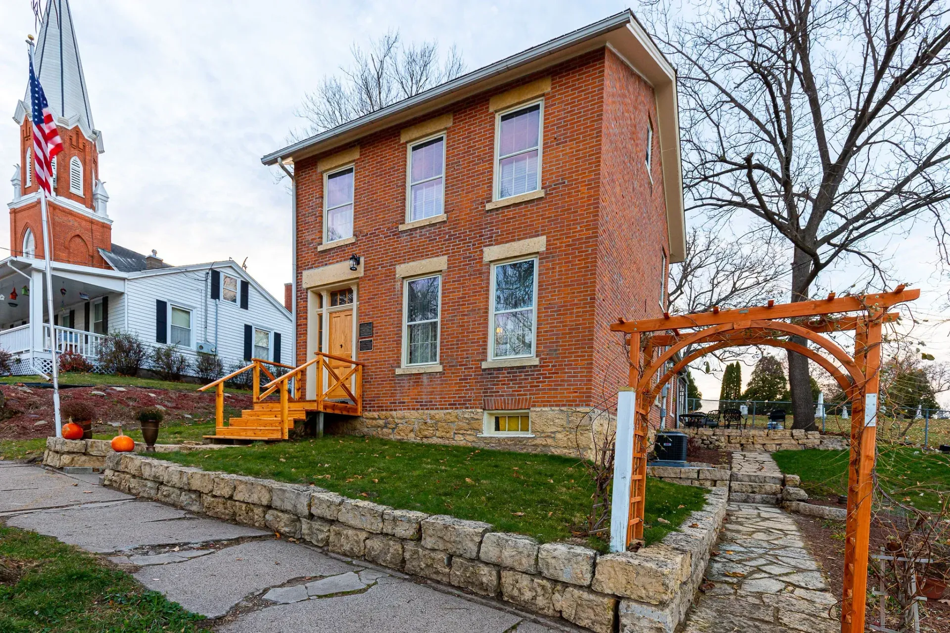 A two-story brick house with a stone foundation and small wooden stairs, next to a church with a tall steeple.