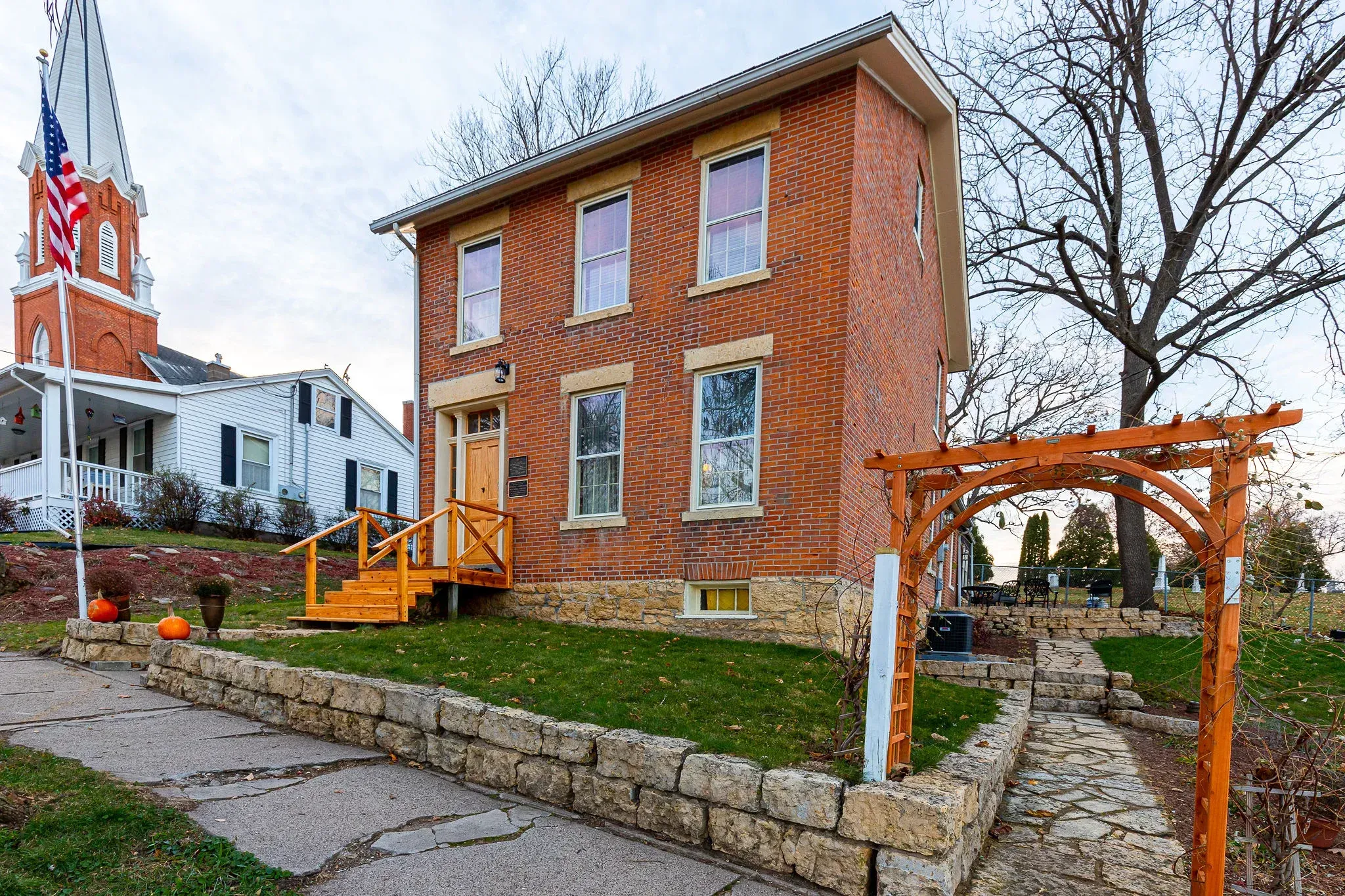A two-story brick house with a stone foundation and small wooden stairs, next to a church with a tall steeple.