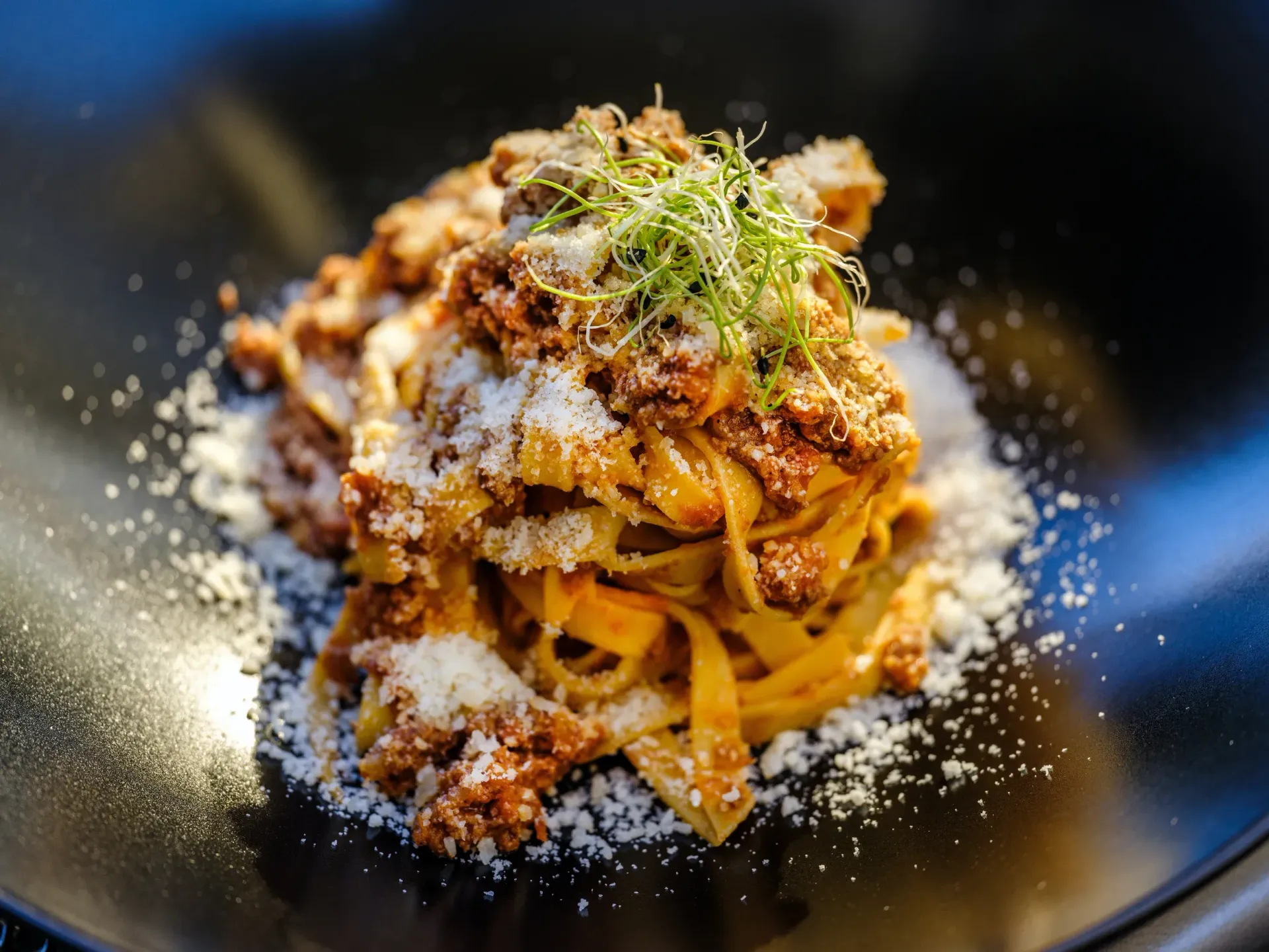 A mound of tagliatelle with meat sauce, topped with grated cheese and microgreens, served in a black bowl.
