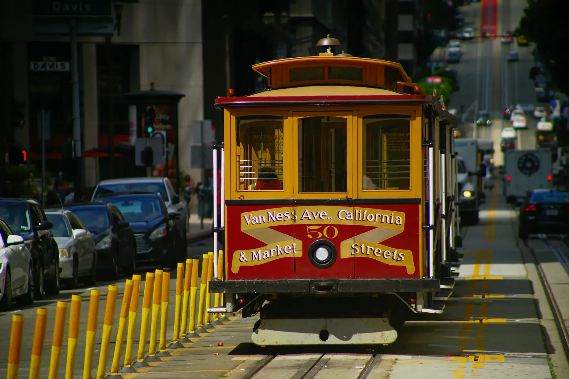 A red and yellow San Francisco cable car travels down a city street lined with yellow bollards and parked cars.
