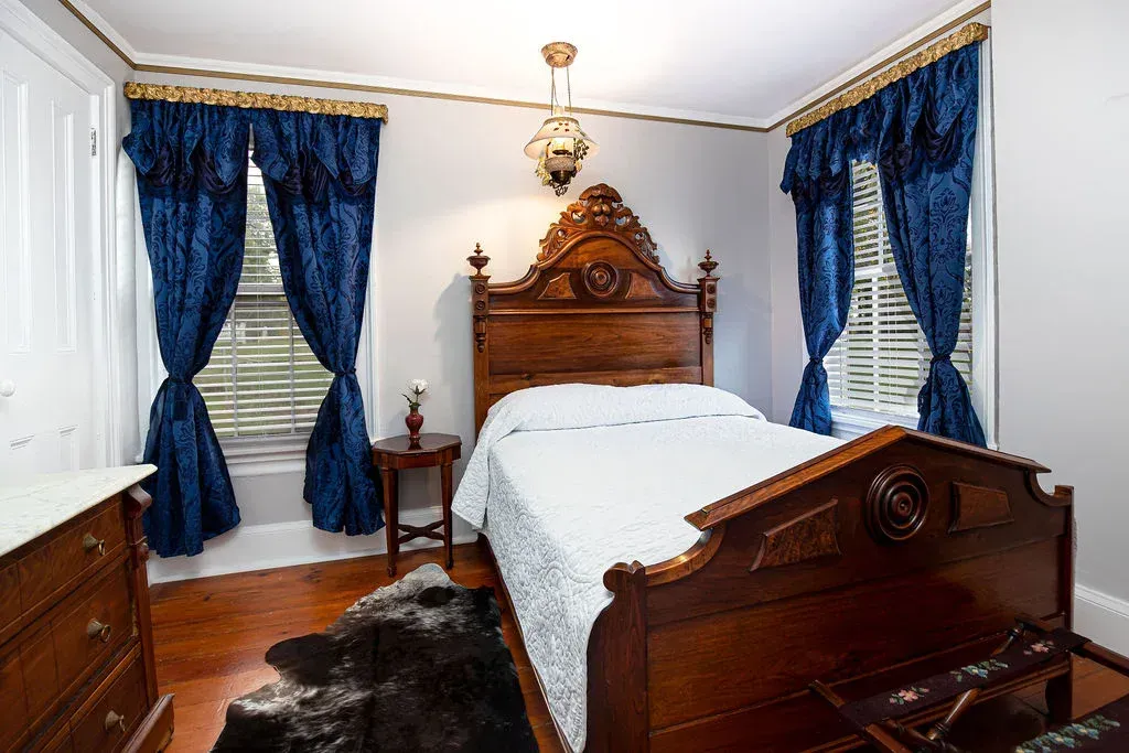 A bedroom with a dark wood carved bed frame, white bedding, blue curtains, a brown wooden side table, and a rug.