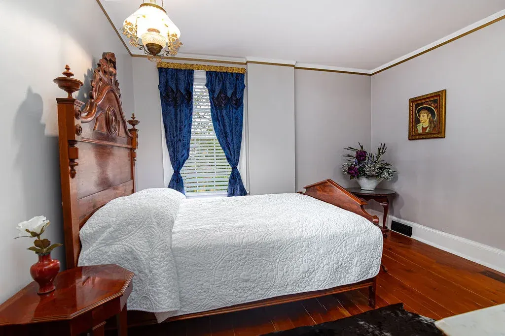 A bedroom with a large carved wooden bed frame, blue drapes, a patterned bedspread, and a small side table with flowers.