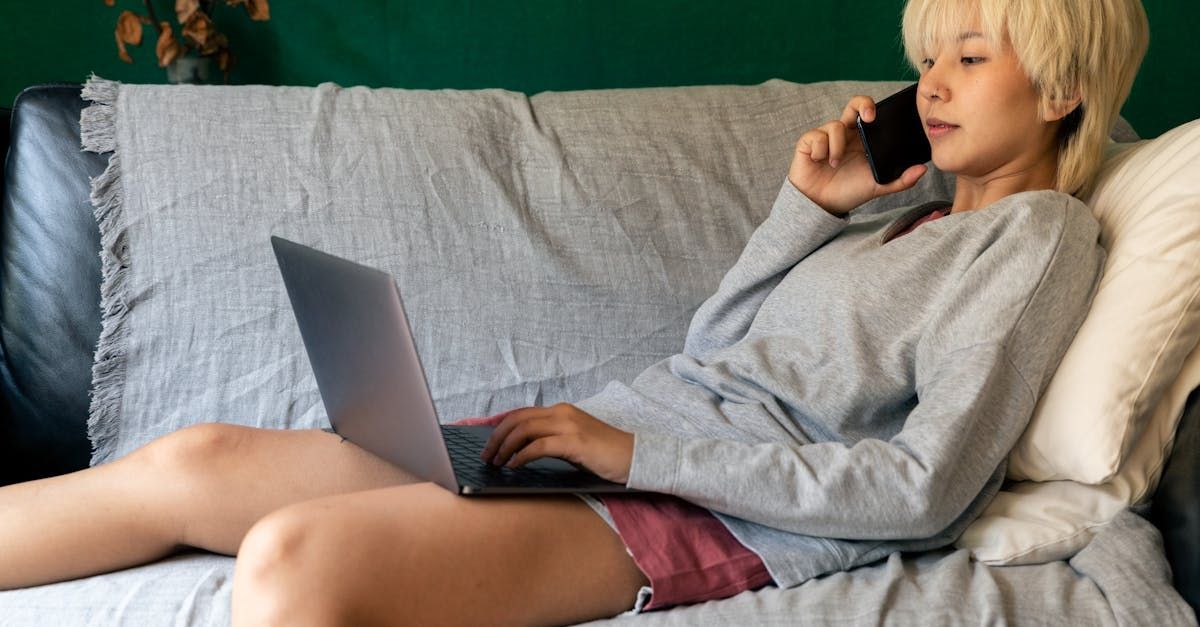 A woman is sitting on a couch using a laptop and talking on a cell phone.