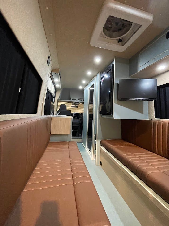 Modern train or bus interior with brown bench seating, overhead lights, and a mounted TV screen