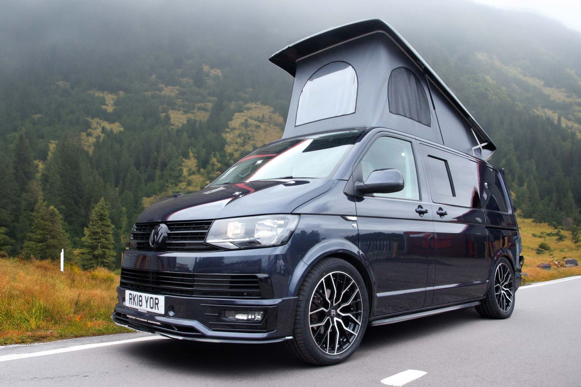 Black camper van with pop-up roof parked on a mountain road beside misty forested slopes