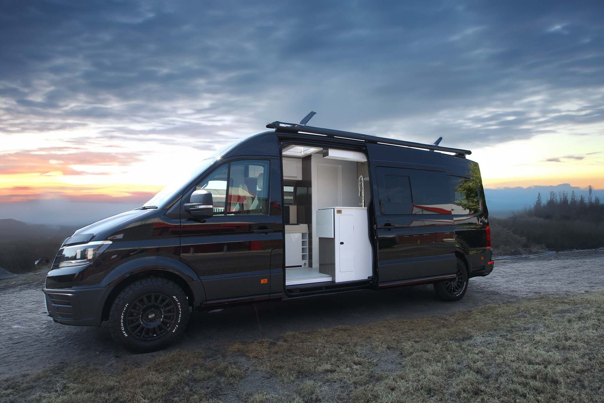 Black camper van parked on gravel at sunset with its sliding door open, revealing a white interior.
