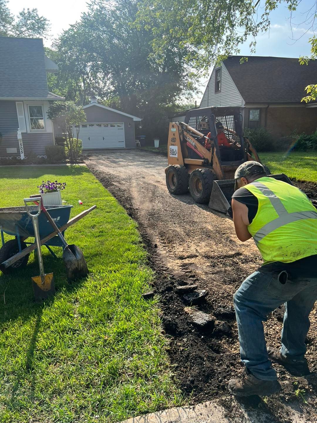 Worker in neon vest using skid steer to spread dirt along a driveway beside a lawn and houses