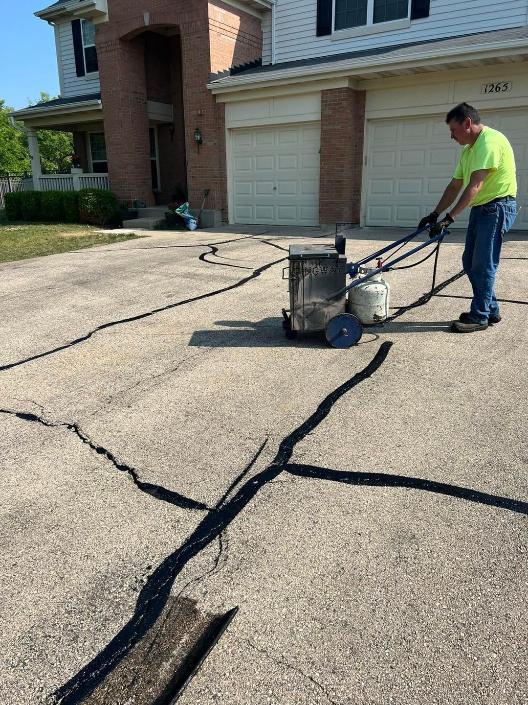 Worker sealing cracks in a driveway with a machine near a suburban garage
