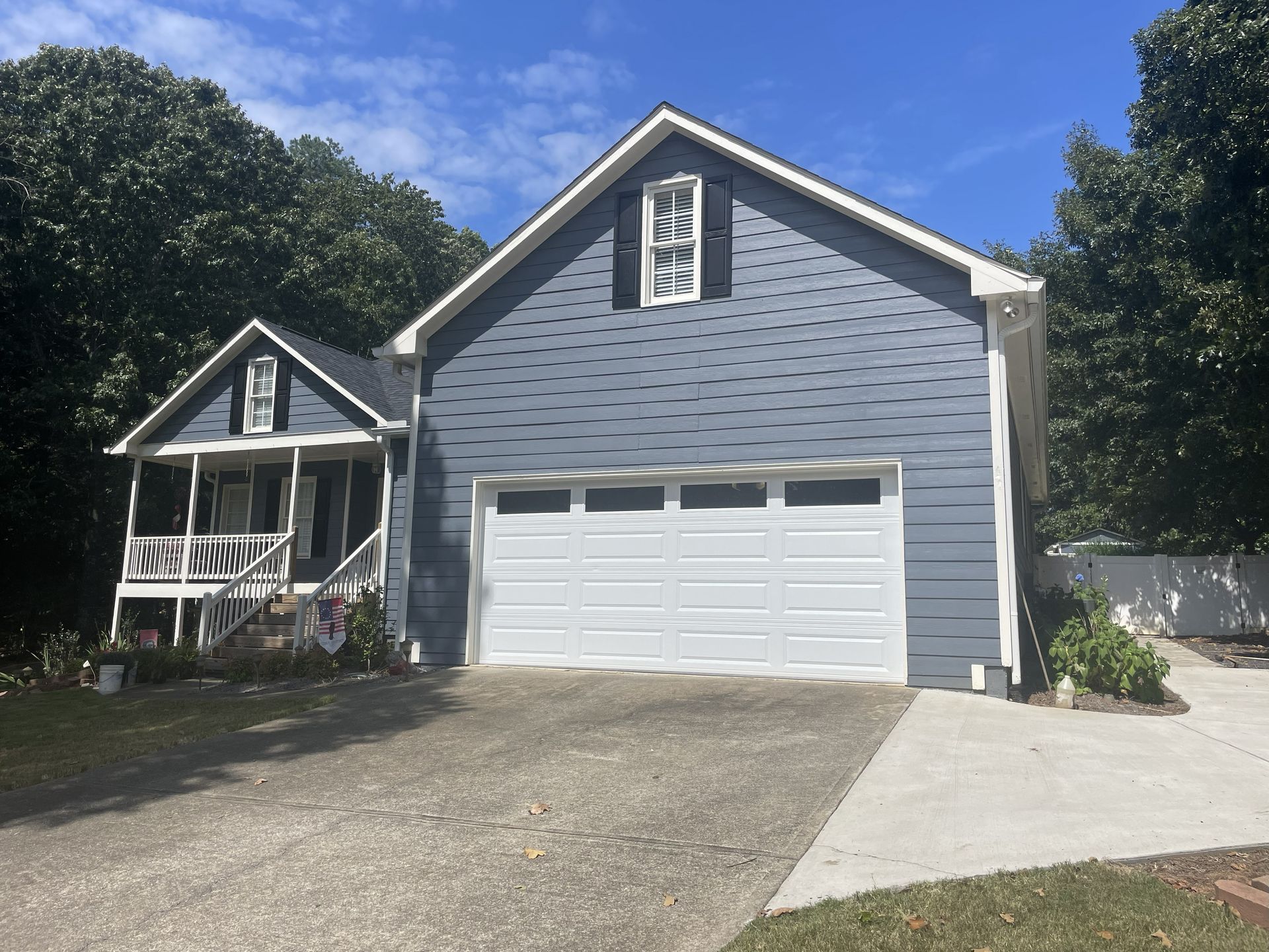 Blue-sided house with a white garage door, small porch, and a driveway, set against a backdrop of trees and blue sky.