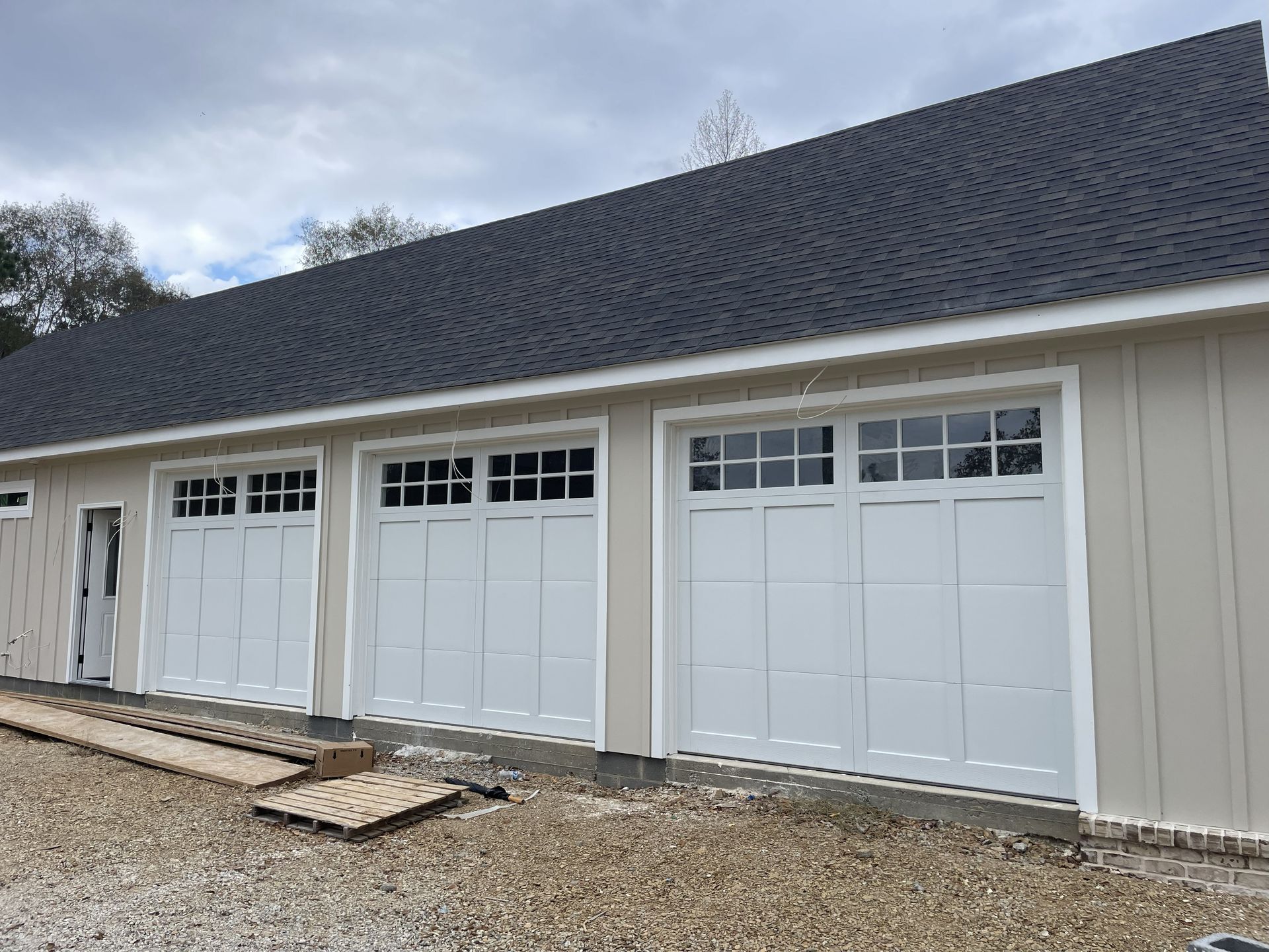 White garage with three doors, tan siding, and a dark roof under a cloudy sky.