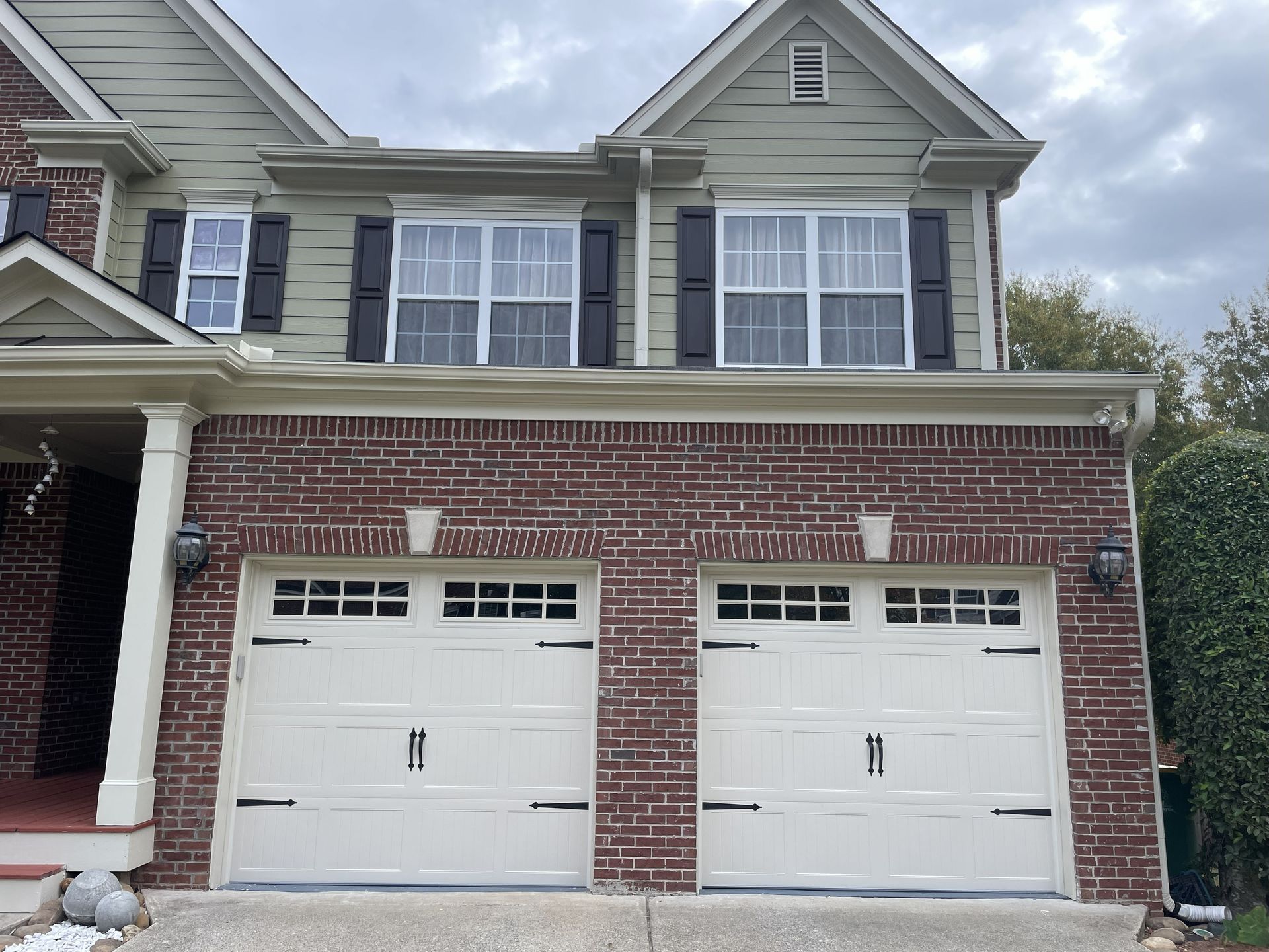 Two-story house with brick base and green siding. Two white garage doors with black accents. Cloudy sky.