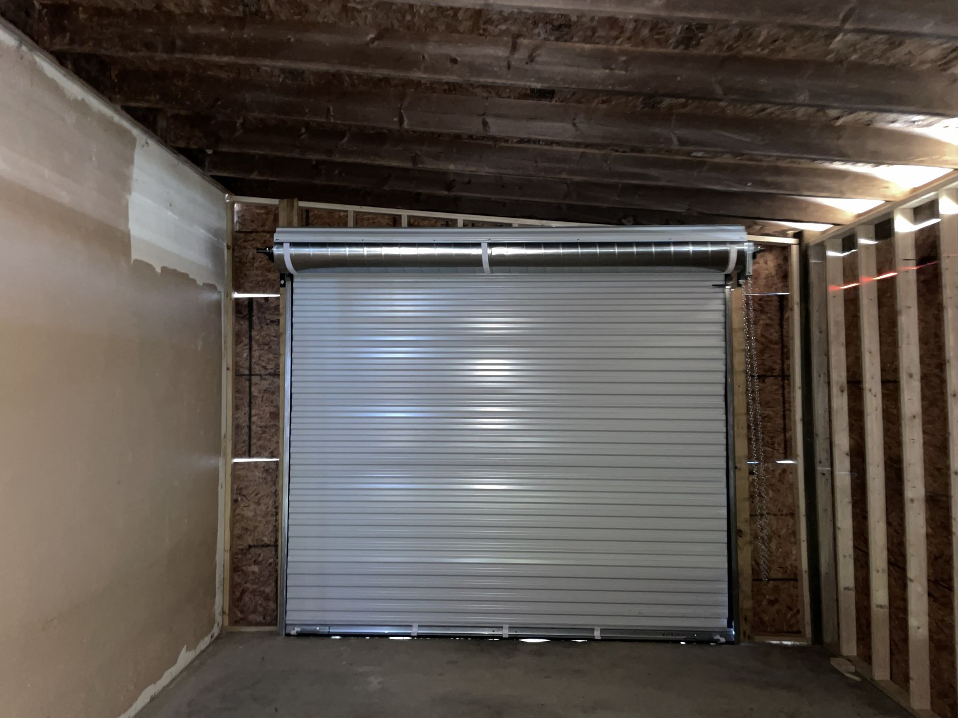 Gray roll-up garage door installed in a framed opening, inside a room with unfinished walls and ceiling.