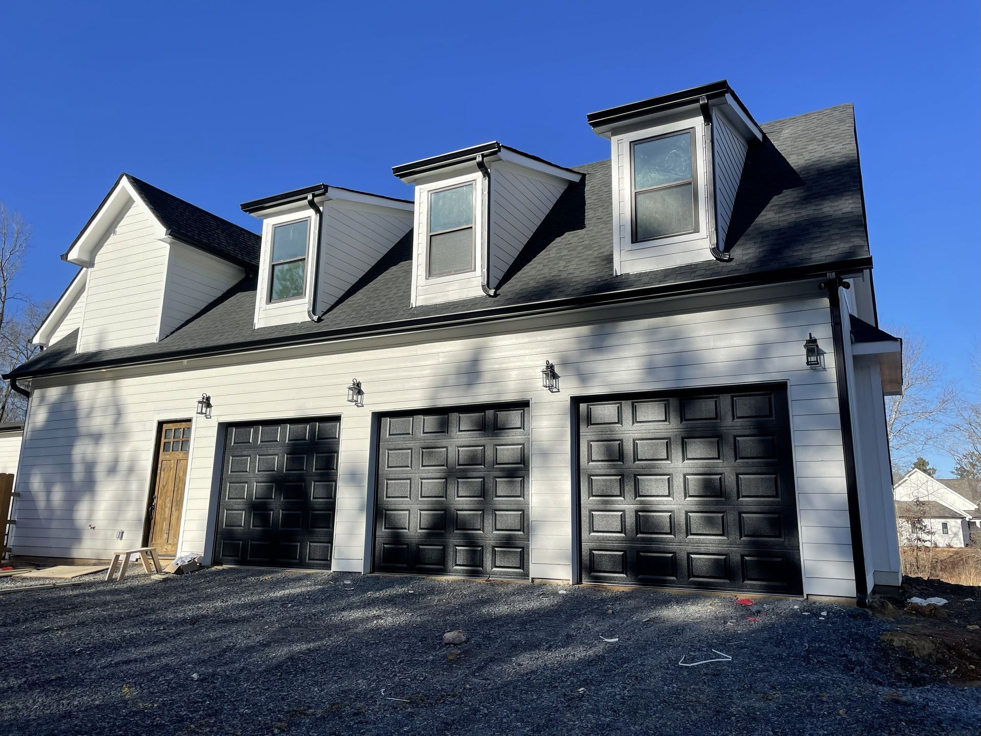 Three-car garage with black doors and dormers, white siding, and a gravel driveway under a blue sky.