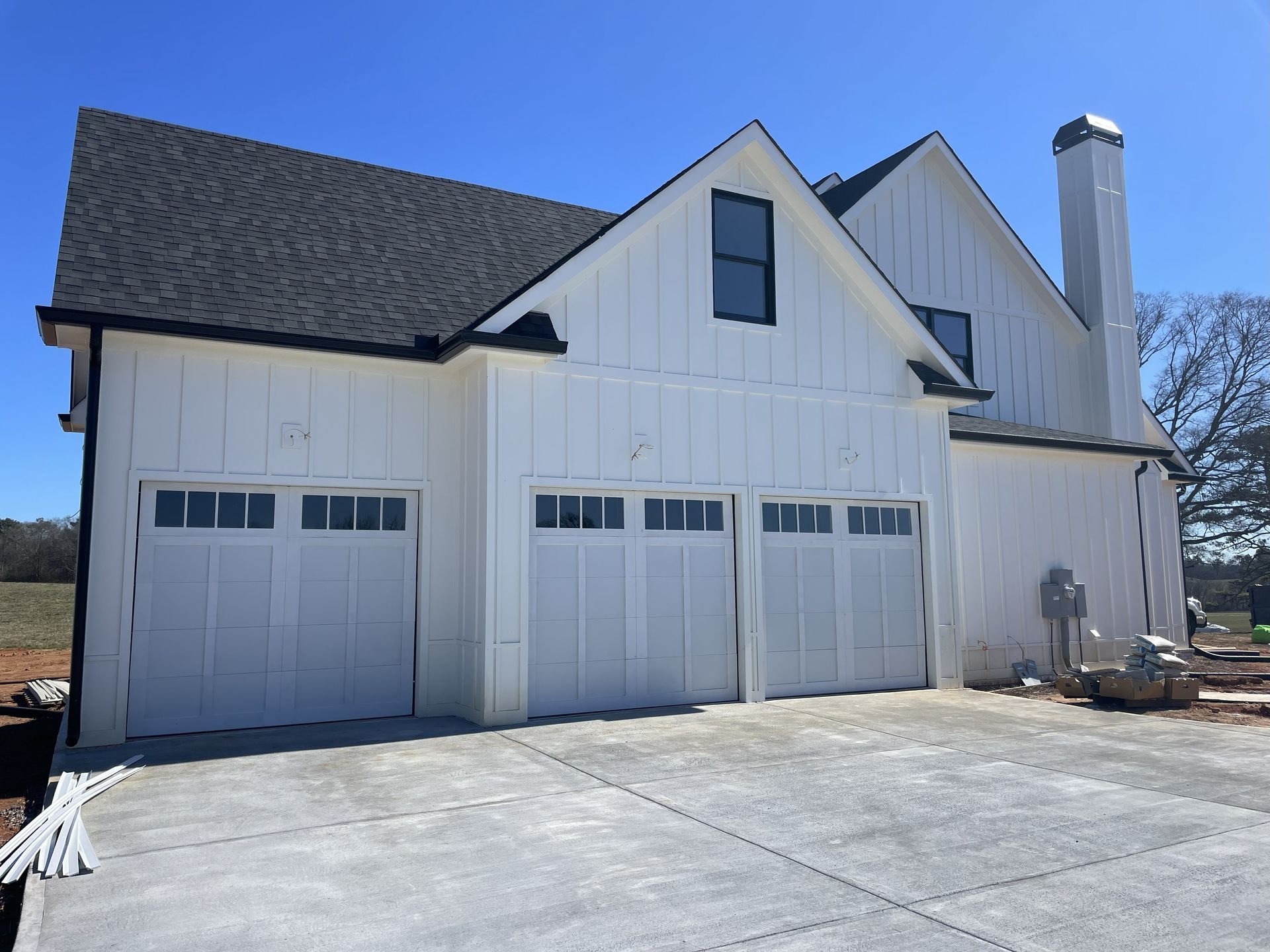 White farmhouse with three-car garage, black roof, and concrete driveway under a blue sky.