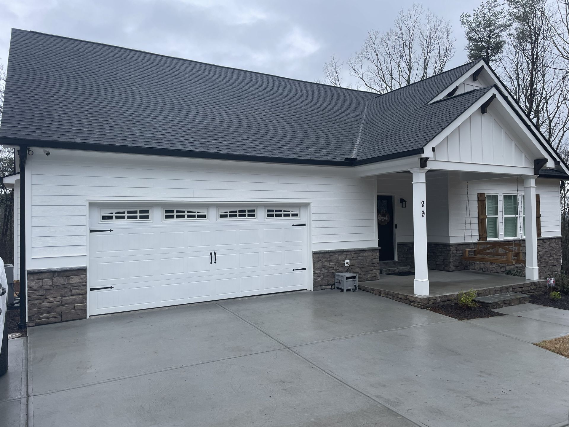 White house with a dark gray roof and a white garage door. Porch with stone accents. Cloudy day.