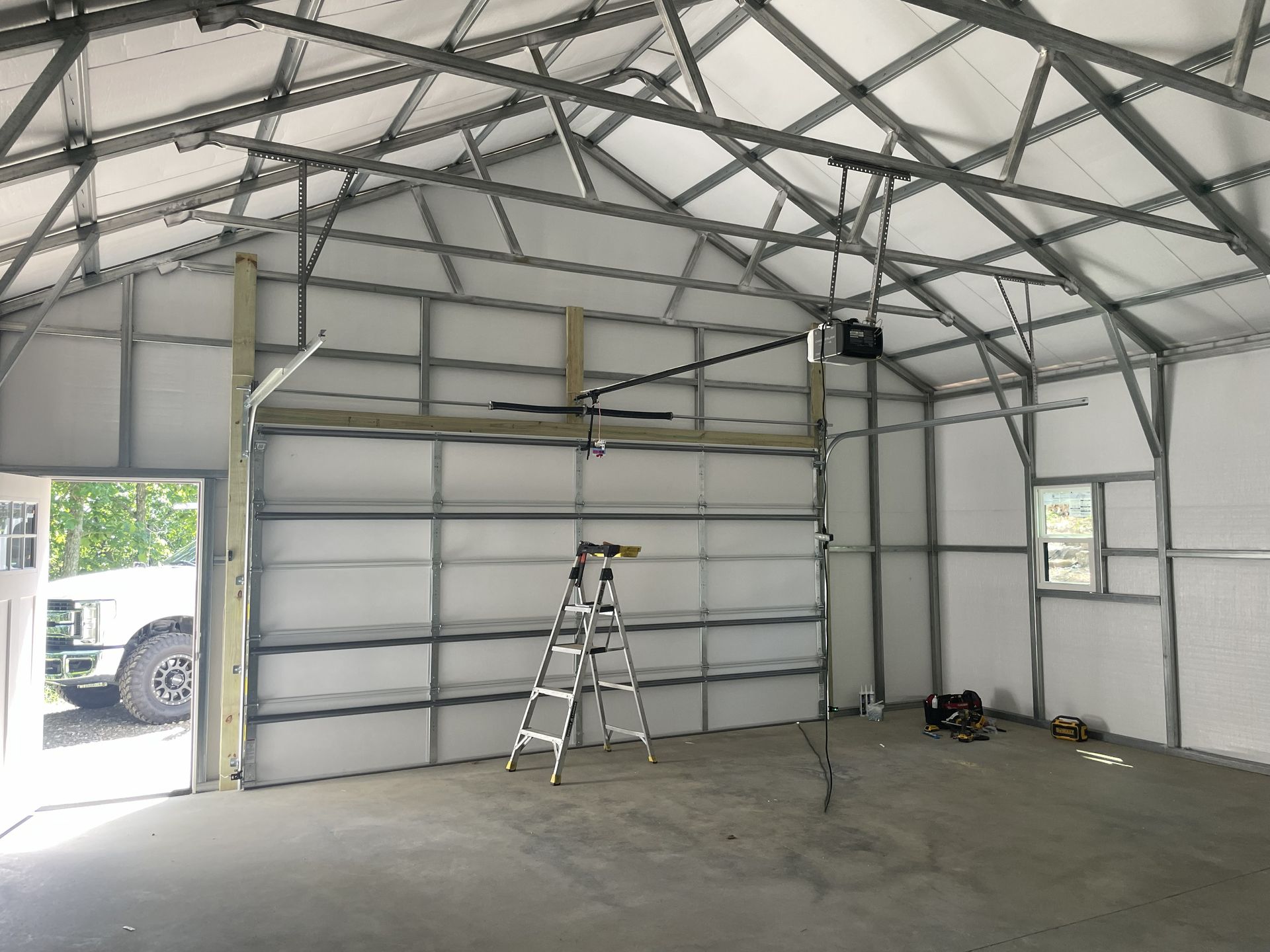 Interior view of a new metal building with a garage door and concrete floor; a ladder and tools are inside.
