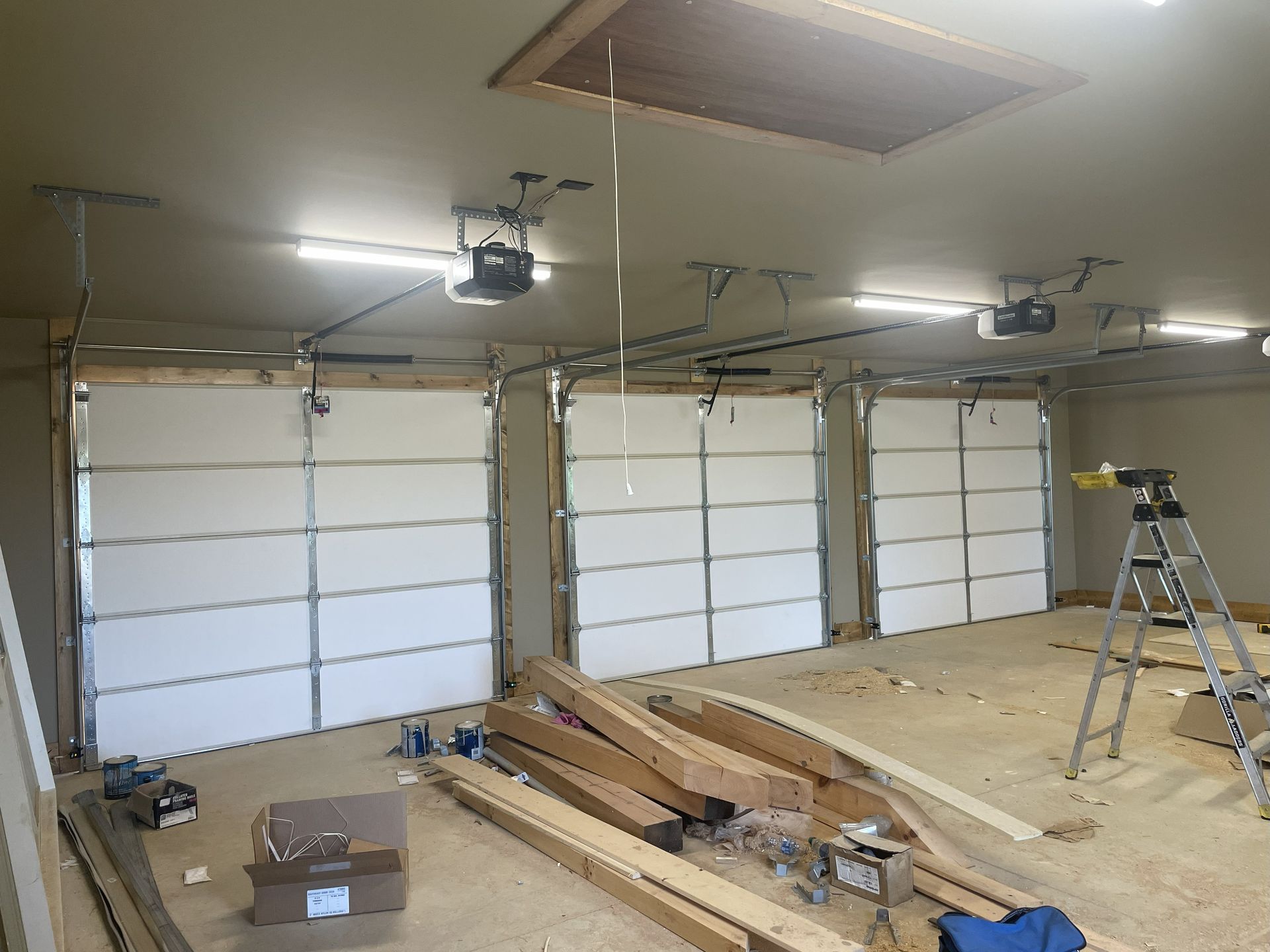 Three white garage doors inside a building, each with an opener. Construction materials are on the floor.