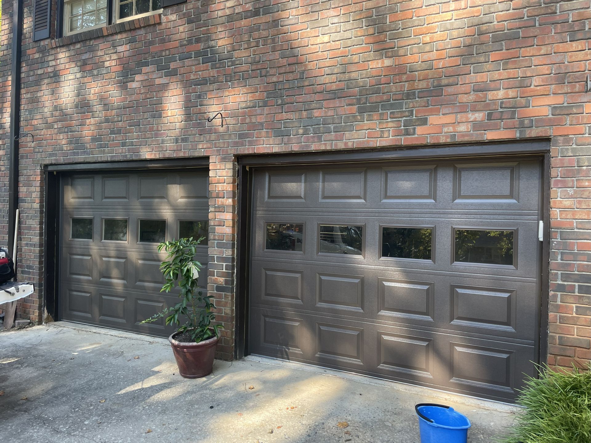 Two brown garage doors on a brick building with a potted plant in between.