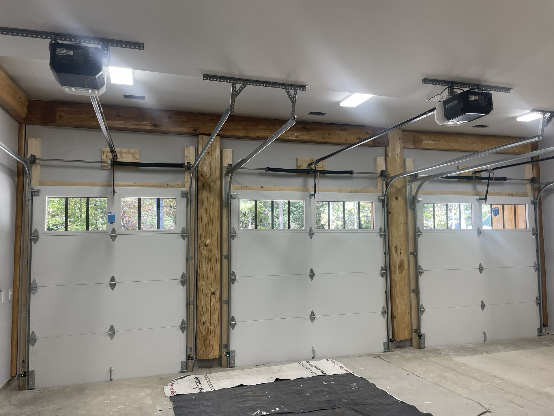 Three white garage doors inside a garage with automatic openers and exposed wood beams.