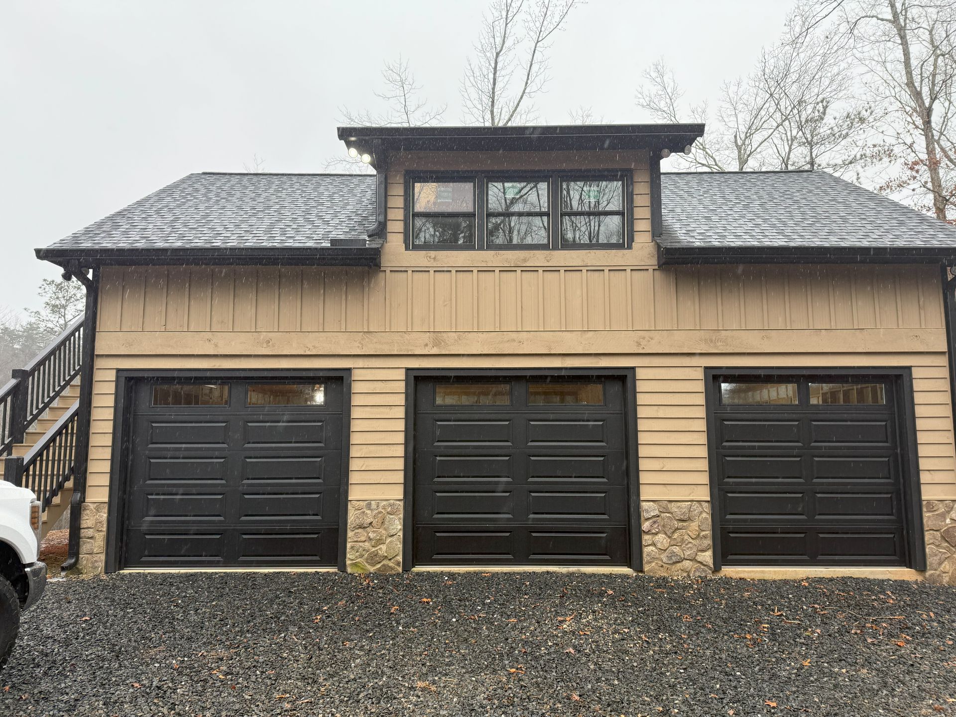 Three-bay garage with black doors and stone accents, beige siding, and an upper window under gray roof.